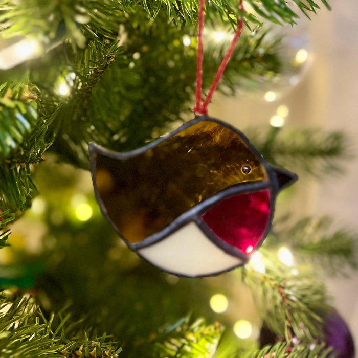 A stained glass bird ornament hanging from a red string on a Christmas tree, with green pine needles and warm white bokeh lights in the background.