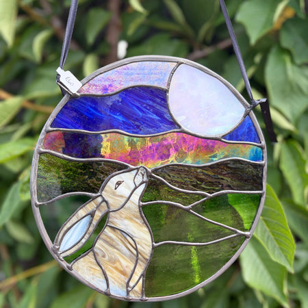 A circular stained glass hanging ornament featuring a rabbit in a field under a blue sky and moon, suspended by a black cord against a background of green leaves.