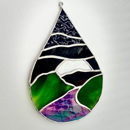 A teardrop-shaped stained glass hanging ornament depicting a landscape with green hills, a purple valley, and a dark blue sky, set against a plain white wall.