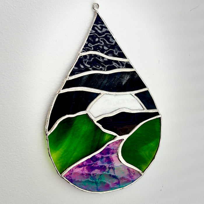 A teardrop-shaped stained glass hanging ornament depicting a landscape with green hills, a purple valley, and a dark blue sky, set against a plain white wall.