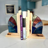 Two handmade stained glass bookends made of patterned blue, green, and brown glass panels on a light wood base, holding up three books on a white marble countertop with a blurred kitchen in the background.