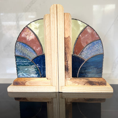 A pair of decorative wooden bookends with stained-glass-style arched panels in blue, brown, beige, and green, supported by light natural wood posts on a reflective dark surface.