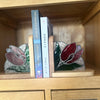 Stained glass tulip bookends on a wooden shelf, holding two books. The bookends feature red and green stained glass tulips with clear glass stems and bases, set against a light oak wooden cabinet.