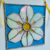 A stained glass hanging ornament shaped like a flower, featuring iridescent white petals, a yellow center, and a bright blue background, suspended by yellow ribbons against a plain wall.