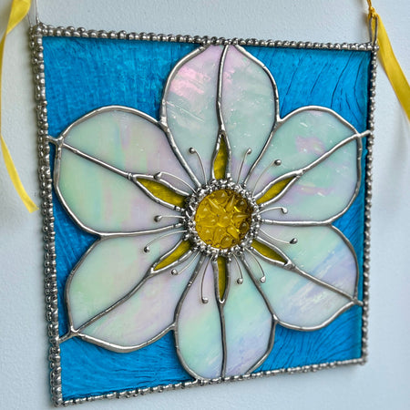 A stained glass hanging ornament shaped like a flower, featuring iridescent white petals, a yellow center, and a bright blue background, suspended by yellow ribbons against a plain wall.