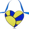 A stained glass heart ornament with a blue ribbon hanger, featuring textured yellow-green and dark blue glass panels separated by dark metal lines, resting on a gray textured surface.
