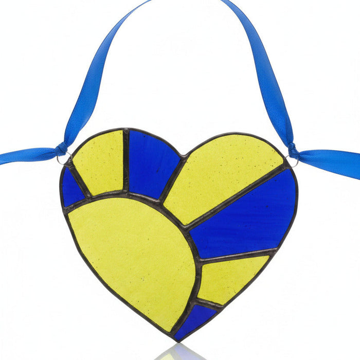 A stained glass heart ornament with a blue ribbon hanger, featuring textured yellow-green and dark blue glass panels separated by dark metal lines, resting on a gray textured surface.