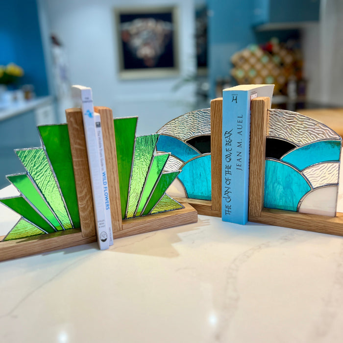 Two decorative bookends with a stained-glass fan design in green, blue, and white, mounted on a light wood base and holding books on a white marble countertop.