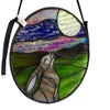 An oval stained glass art piece featuring a hare, a sun, and flowing bands of blue, purple, green, and black, shown hanging against a plain white wall.