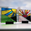 Three small stained glass art panels displayed on black stands on a white surface, with a softly blurred cloudy sky in the background. The left panel shows blue, yellow, green, and black geometric shapes; the center panel shows a multicolor sunburst with pink, orange, yellow, red, and white; the right panel shows a night landscape with deep blue, brown, and black.