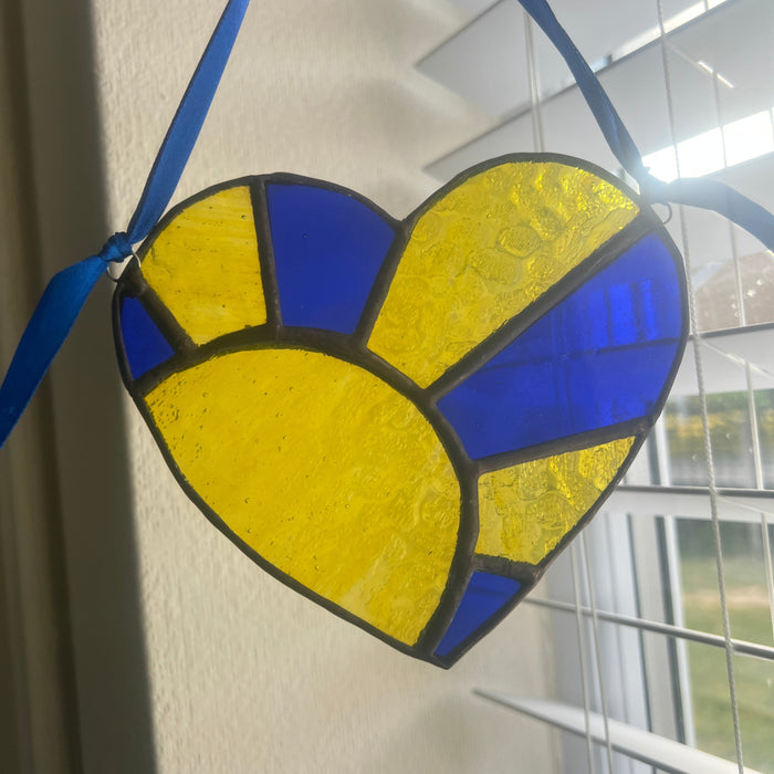 A heart-shaped stained glass ornament with blue and yellow panels, held by a hand in front of a window with blinds.