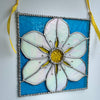 A square stained glass hanging ornament featuring a large white flower with a yellow center on a textured blue background, outlined with silver beaded metal and suspended by a yellow ribbon.