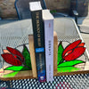 Stained glass bookends with red tulips and green leaves on a wooden base, holding two books on an outdoor patio table.