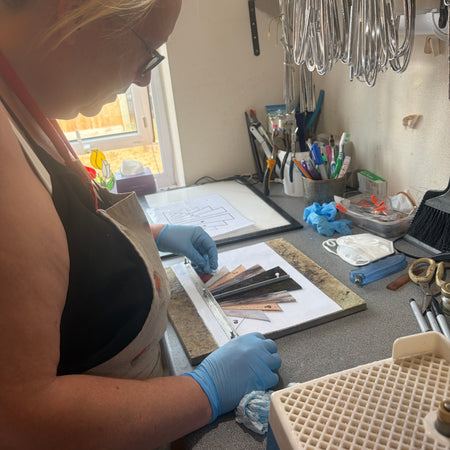 A person wearing a black top, beige apron, and blue gloves works at a craft table, handling small metal components in a ring binder. The workspace contains tools, brushes, hanging metal loops, and other crafting supplies.