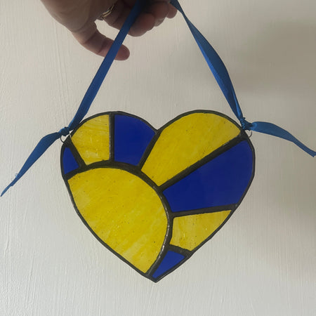 A heart-shaped stained glass ornament with blue and yellow panels and blue hanging ribbons, held by a hand against a plain off-white wall.