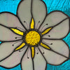 A close-up of a stained glass flower design with pale lavender-white petals, a textured gold center, and dark lead lines on a bright blue background.