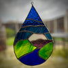 A teardrop-shaped stained glass hanging ornament featuring a landscape design with blue sky, green hills, and purple mountains, suspended by a metal ring against a blurred outdoor background.
