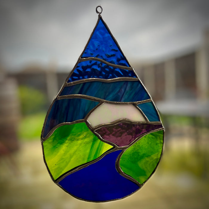 A teardrop-shaped stained glass hanging ornament featuring a landscape design with blue sky, green hills, and purple mountains, suspended by a metal ring against a blurred outdoor background.