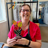 A smiling woman wearing glasses and a red shirt holds a small potted stained glass flower arrangement with yellow, orange, and green leaves in a workshop or studio setting.