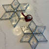Two six-pointed star Christmas ornaments made of textured clear glass with silver metal borders and a small red hanging ribbon, displayed on a light marble surface.