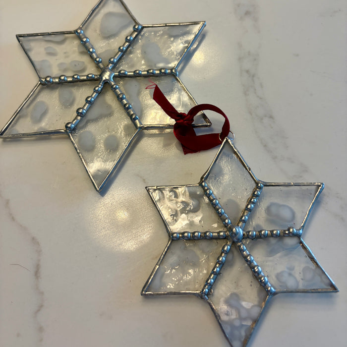 Two six-pointed star Christmas ornaments made of textured clear glass with silver metal borders and a small red hanging ribbon, displayed on a light marble surface.