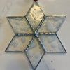 A handmade six-pointed star glass ornament with a hammered glass texture, silver metal borders, and a red hanging ribbon, shown on a white marble surface.