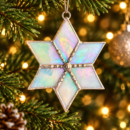 A six-pointed star Christmas ornament with iridescent opal-like panels and silver-toned metal trim, hanging in front of a blurred Christmas tree with warm lights.