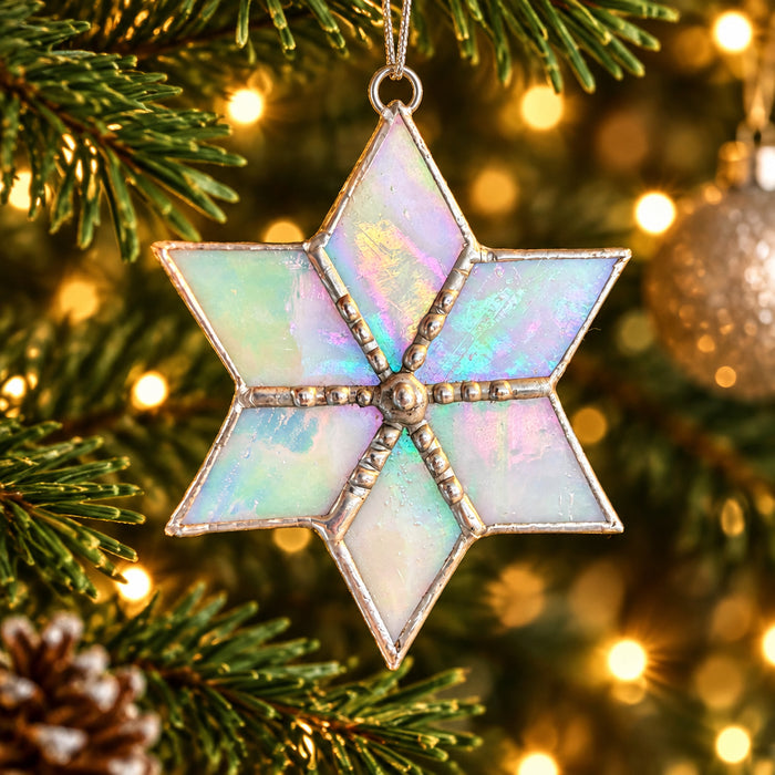 A six-pointed star Christmas ornament with iridescent opal-like panels and silver-toned metal trim, hanging in front of a blurred Christmas tree with warm lights.