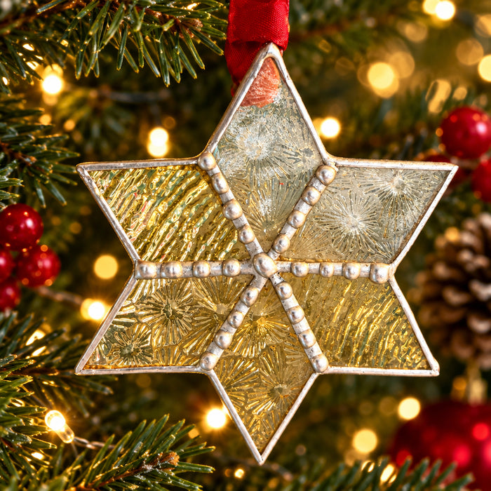 A six-pointed star Christmas ornament hanging on a lit Christmas tree. The ornament has a textured gold and frosted silver design divided by silver beaded lines, with a red hanging ribbon.