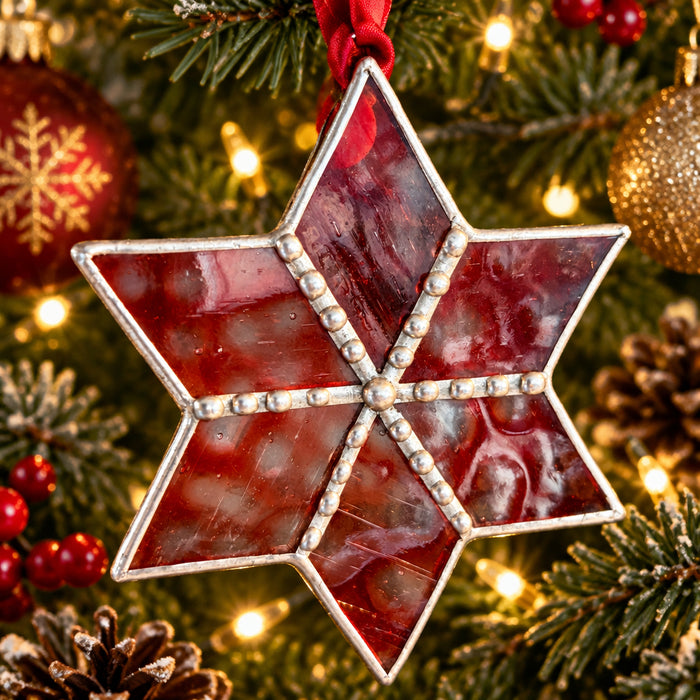 A six-pointed star-shaped stained glass ornament with a red and brown marbled pattern, held together by silver-colored beaded lines, hanging from a red ribbon in a blurred kitchen setting.