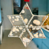 A six-pointed star-shaped hanging ornament made of clear resin with embedded white heart-shaped pieces and silver beaded trim, shown hanging indoors against a blurred blue kitchen background.