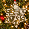 A six-pointed star-shaped glass ornament with a silver-toned metal border, decorated with white and gray marbled resin inclusions and small metallic beads along the seams, hanging against a blurred kitchen background.
