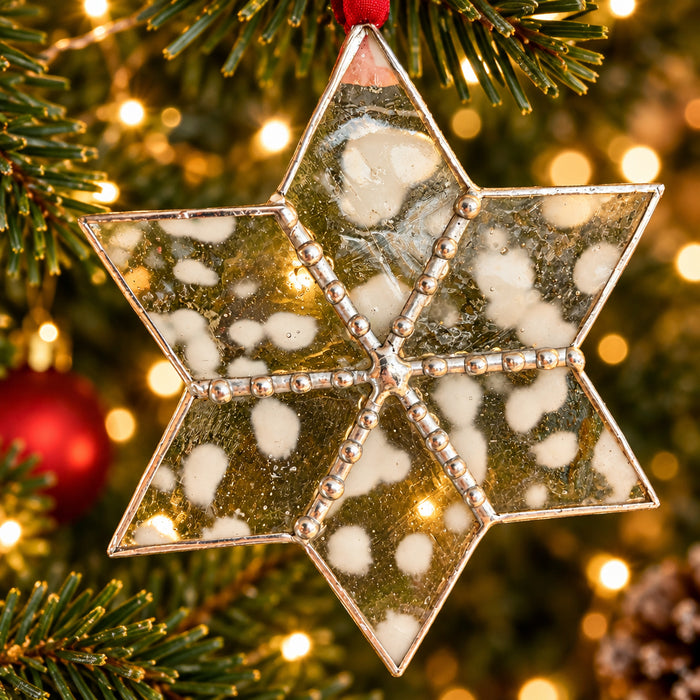 A six-pointed star-shaped glass ornament with a silver-toned metal border, decorated with white and gray marbled resin inclusions and small metallic beads along the seams, hanging against a blurred kitchen background.