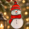 A hand holds a snowman-shaped hanging stained glass ornament with an orange hat and scarf, white body, and silver metal edging, set against a warm blurred holiday background with lights and pine branches.