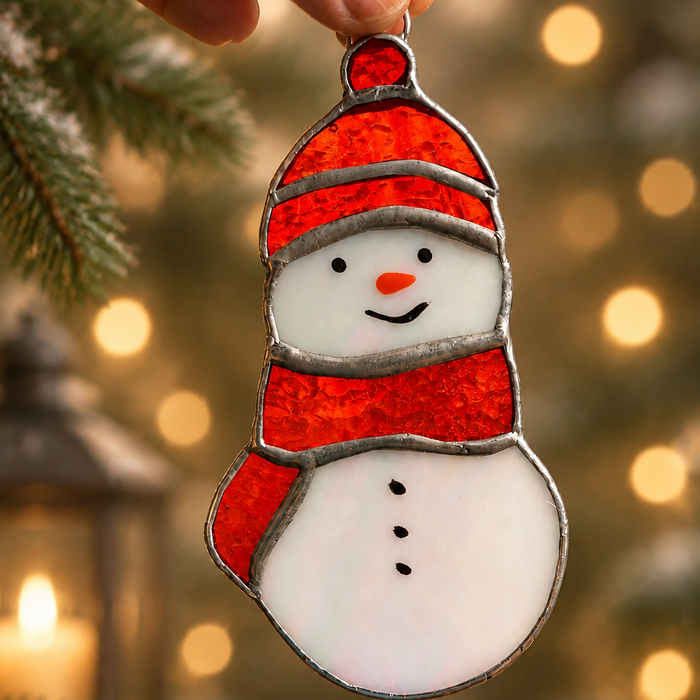 A hand holds a snowman-shaped hanging stained glass ornament with an orange hat and scarf, white body, and silver metal edging, set against a warm blurred holiday background with lights and pine branches.