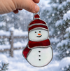 A hand holds a stained glass snowman ornament with a red hat and scarf against a snowy outdoor background.