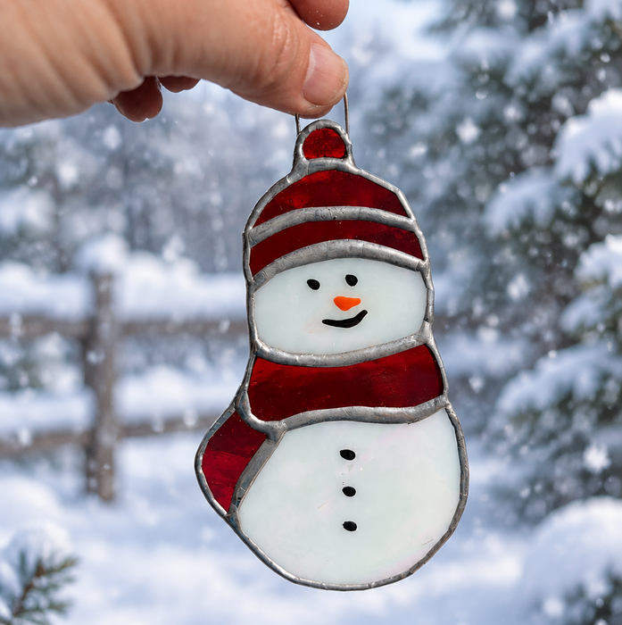 A hand holds a stained glass snowman ornament with a red hat and scarf against a snowy outdoor background.