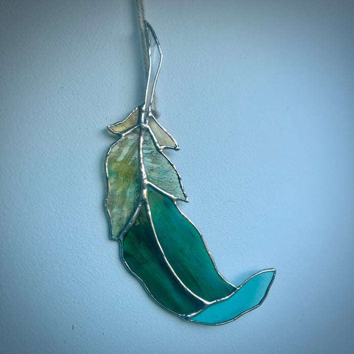 A stained glass feather ornament in shades of green and turquoise, suspended by a cord against a light blue wall.