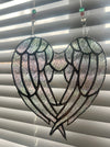 A stained-glass-style angel wings heart suncatcher hanging in front of window blinds. The design features two large wings with textured, iridescent glass panels outlined in black metal, suspended by strings with decorative beads.