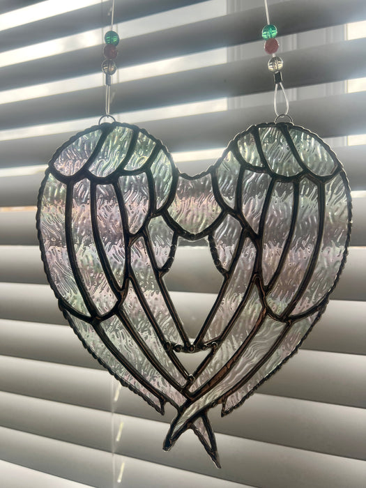 A stained-glass-style angel wings heart suncatcher hanging in front of window blinds. The design features two large wings with textured, iridescent glass panels outlined in black metal, suspended by strings with decorative beads.