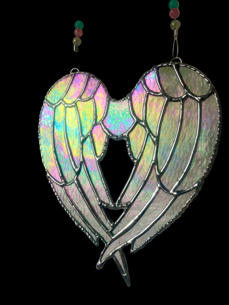 A heart-shaped stained glass suncatcher featuring angel wings with an iridescent, holographic finish and dark silver metal outlines, hanging against a plain light gray wall.