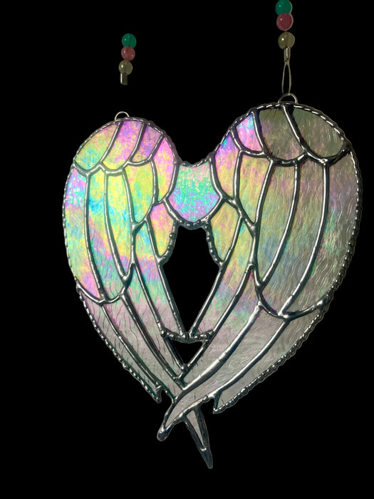 A heart-shaped stained glass suncatcher featuring angel wings with an iridescent, holographic finish and dark silver metal outlines, hanging against a plain light gray wall.