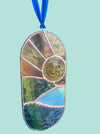 A stained glass suncatcher with a sunburst design, featuring a central circular window and multiple geometric panels in gold, green, blue, and brown tones, suspended by a blue ribbon.