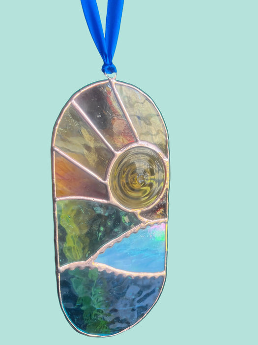 A stained glass suncatcher with a sunburst design, featuring a central circular window and multiple geometric panels in gold, green, blue, and brown tones, suspended by a blue ribbon.