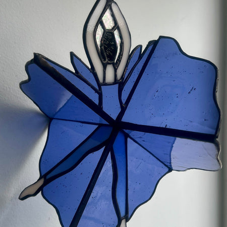 A stained glass suncatcher depicting a ballerina in a blue tutu with black lead lines, hanging against a plain white wall.