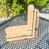 A wooden corner bracket with overlaid dimension arrows and labels showing measurements of approximately 15cm by 13.2cm, placed on a black metal mesh table with green foliage in the background.