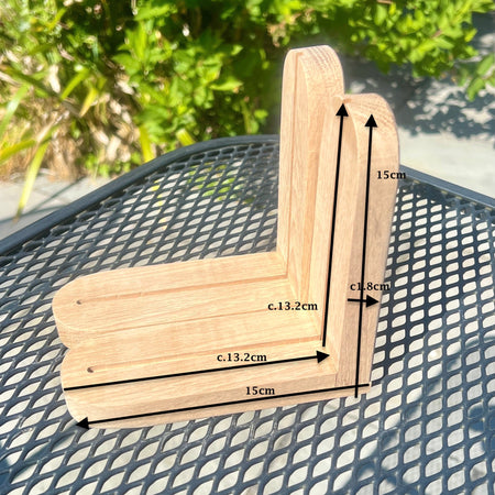 A wooden corner bracket with overlaid dimension arrows and labels showing measurements of approximately 15cm by 13.2cm, placed on a black metal mesh table with green foliage in the background.