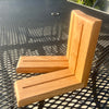 A pair of light oak wood corner brackets with a smooth finish and visible wood grain, resting on a black metal mesh patio table.
