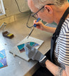 A craftsman wearing a striped shirt and gray work gloves uses a soldering iron to work on a small stained glass piece on a metal workbench.