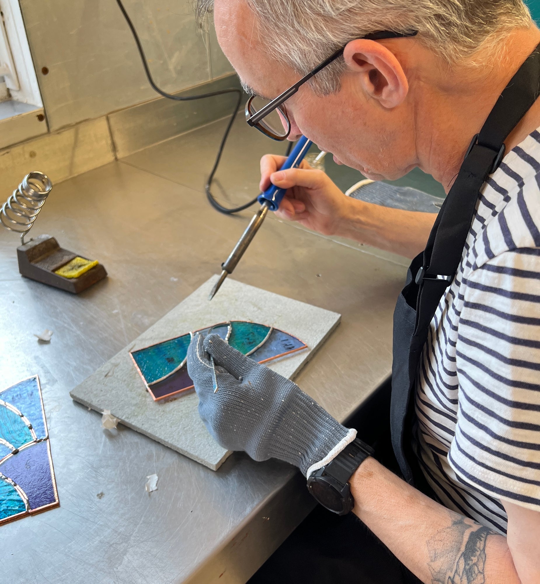 A craftsman wearing a striped shirt and gray work gloves uses a soldering iron to work on a small stained glass piece on a metal workbench.